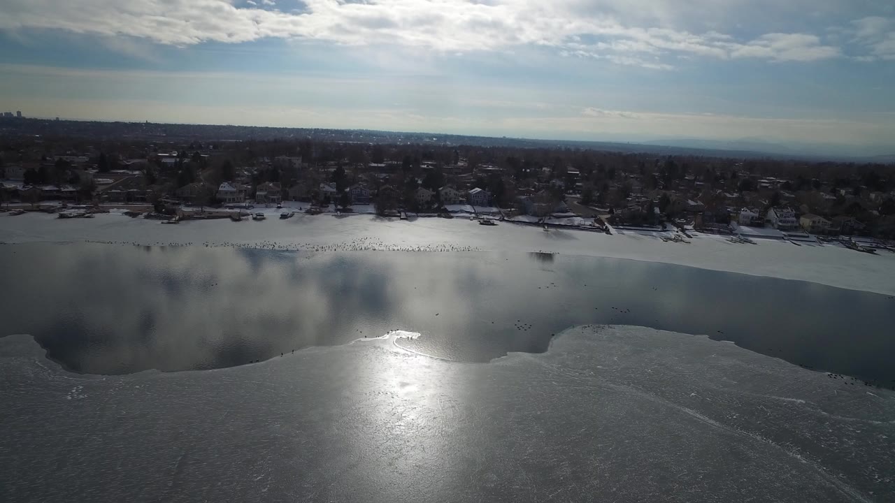 Aerial view of frozen lake beside a residential shoreline under scattered clouds.