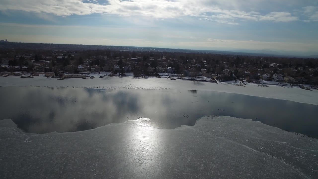 Hidden Lake aerial frame showing a broad icy foreground, a darker reflective water band, distant residential homes, winter trees, and no visible people.