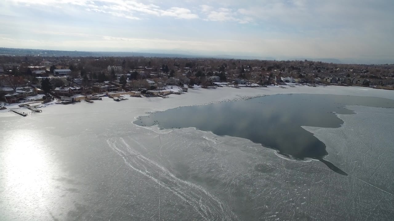 Aerial view of frozen lake and shoreline homes under pale sky.