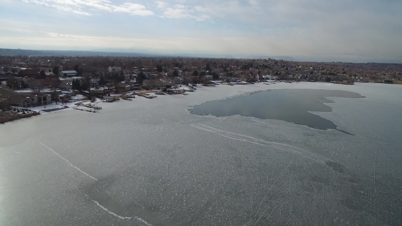Aerial view over a partially frozen lake toward a residential shoreline.