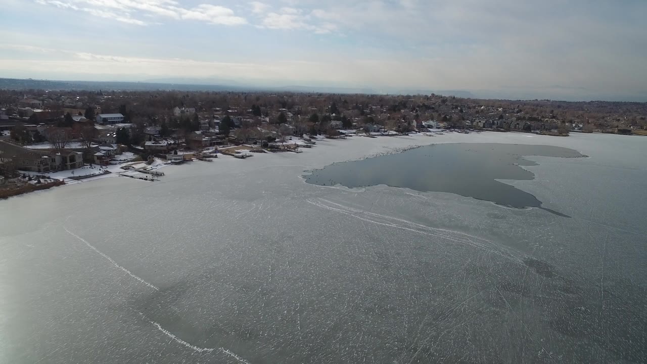 Hidden Lake, Colorado appears from above with textured ice across the foreground, a darker open-water patch by the shore, homes and trees along the bank, and no visible people.