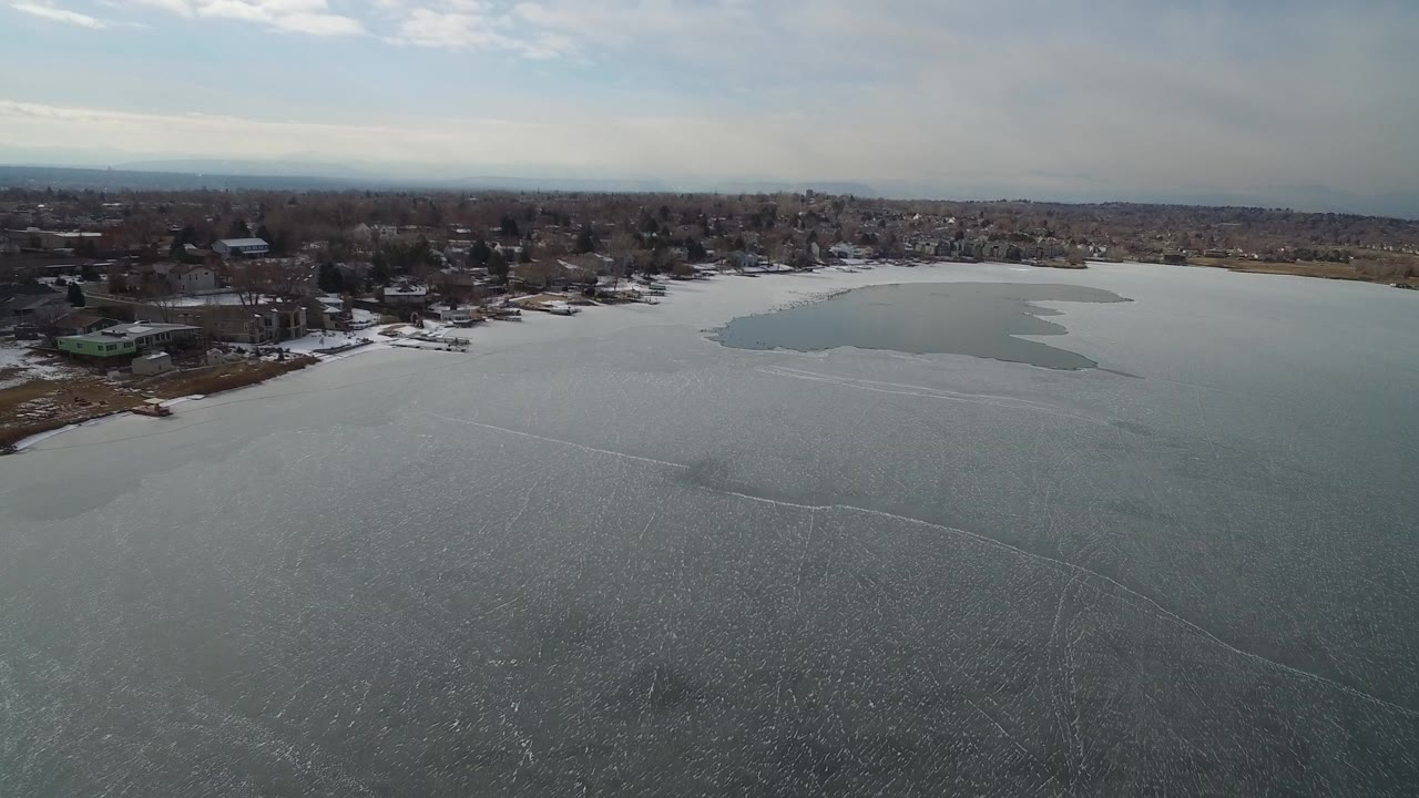 Aerial view over frozen water with shoreline homes and bare trees.