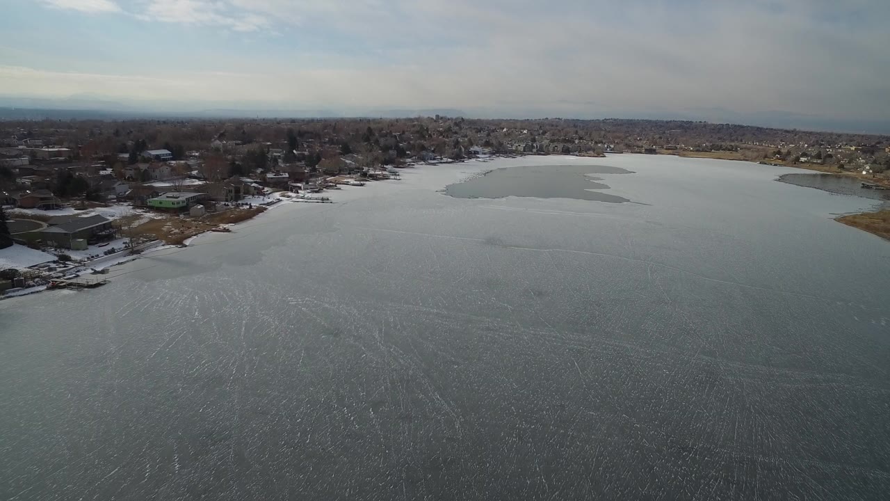 Aerial frame over Hidden Lake, Colorado, showing gray-blue water or thin ice in the foreground, snow along the shoreline, residential homes beyond, and no visible people.