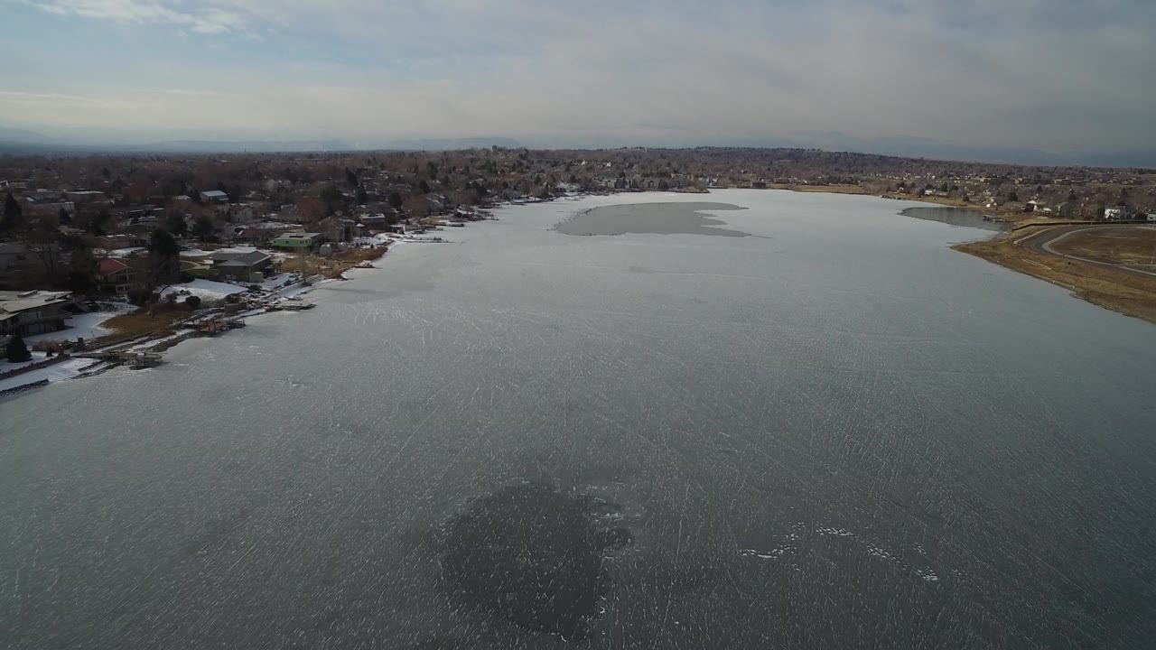 Aerial view across gray frozen water toward shoreline homes and roads.