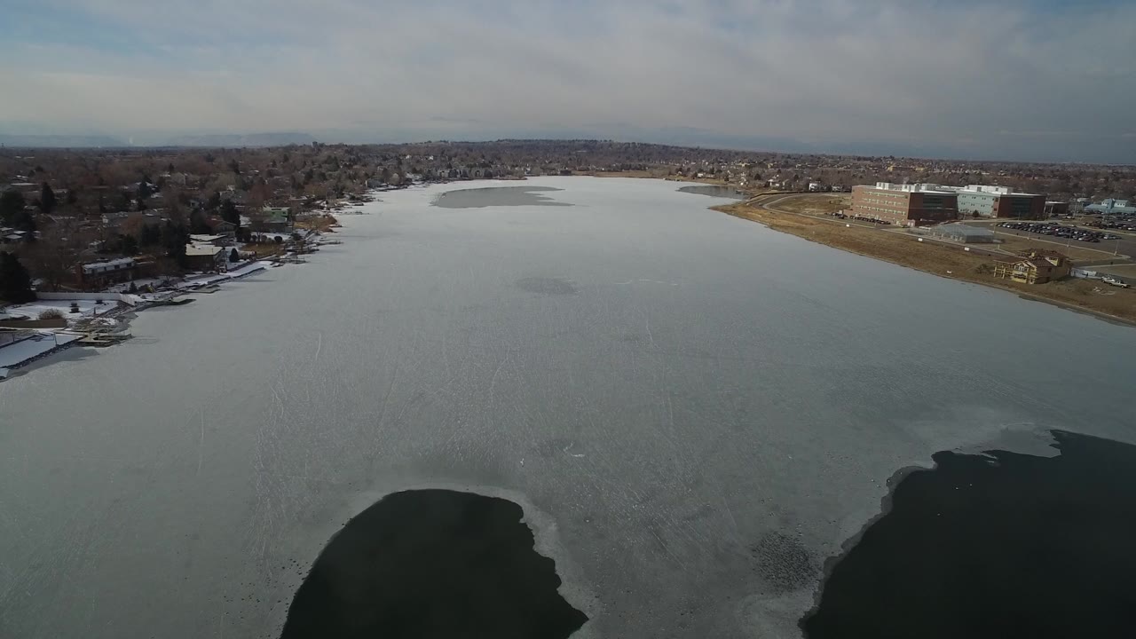 Aerial frame over Hidden Lake, Colorado, showing gray lake ice, dark open-water patches near the foreground, snowy banks, distant homes, and no visible people.