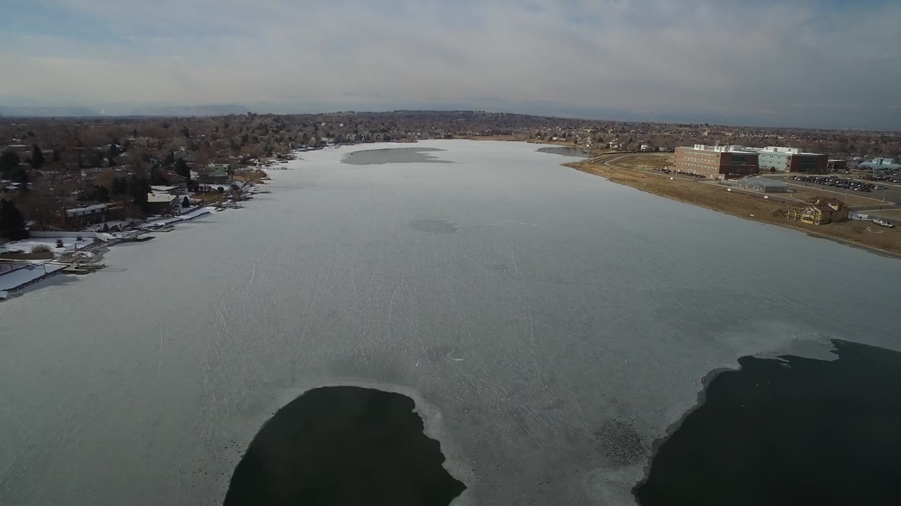 High view of Hidden Lake, Colorado, with a broad gray lake, darker broken ice areas in the lower frame, shoreline houses, larger low buildings, and no visible people.