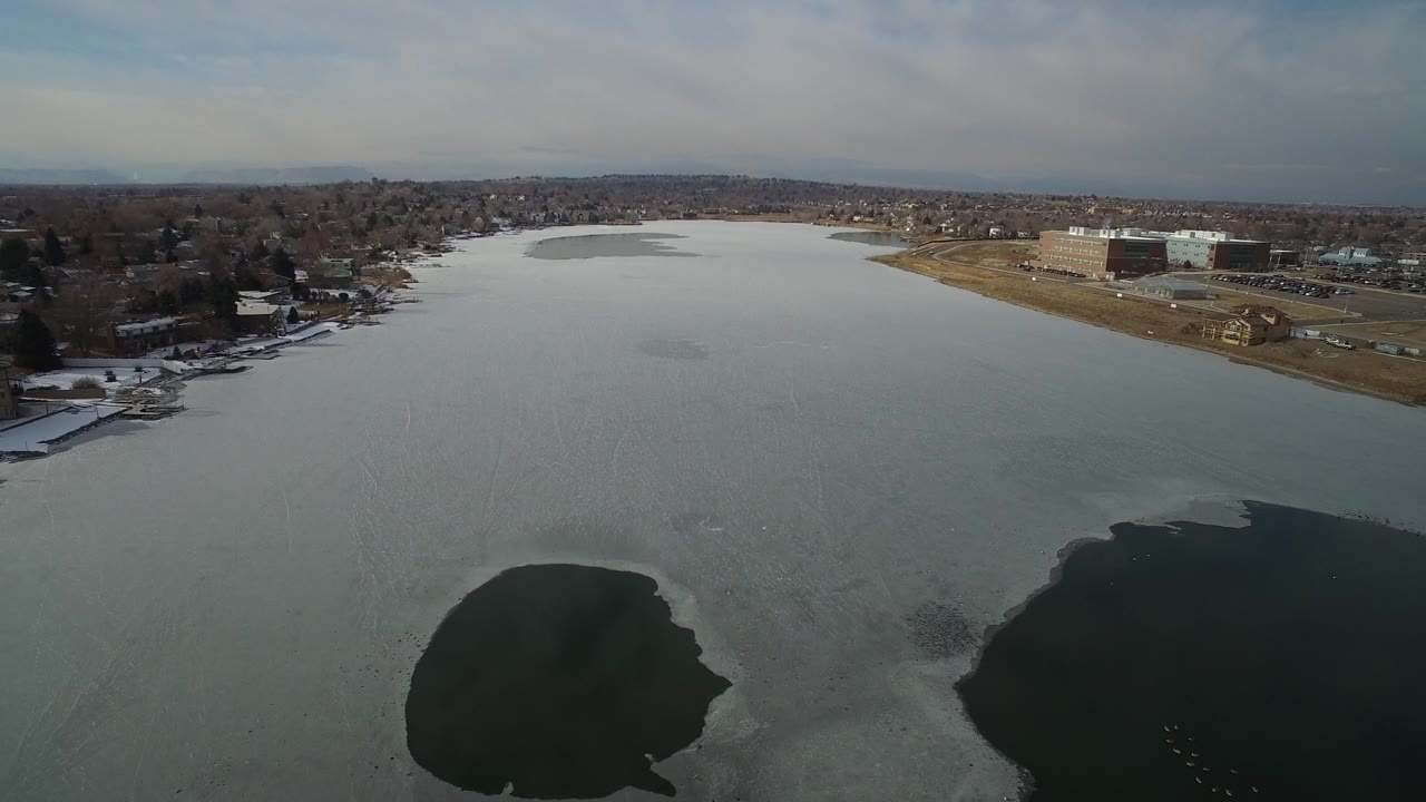 Aerial lake view with suburban shoreline, roads, and larger buildings in the distance.