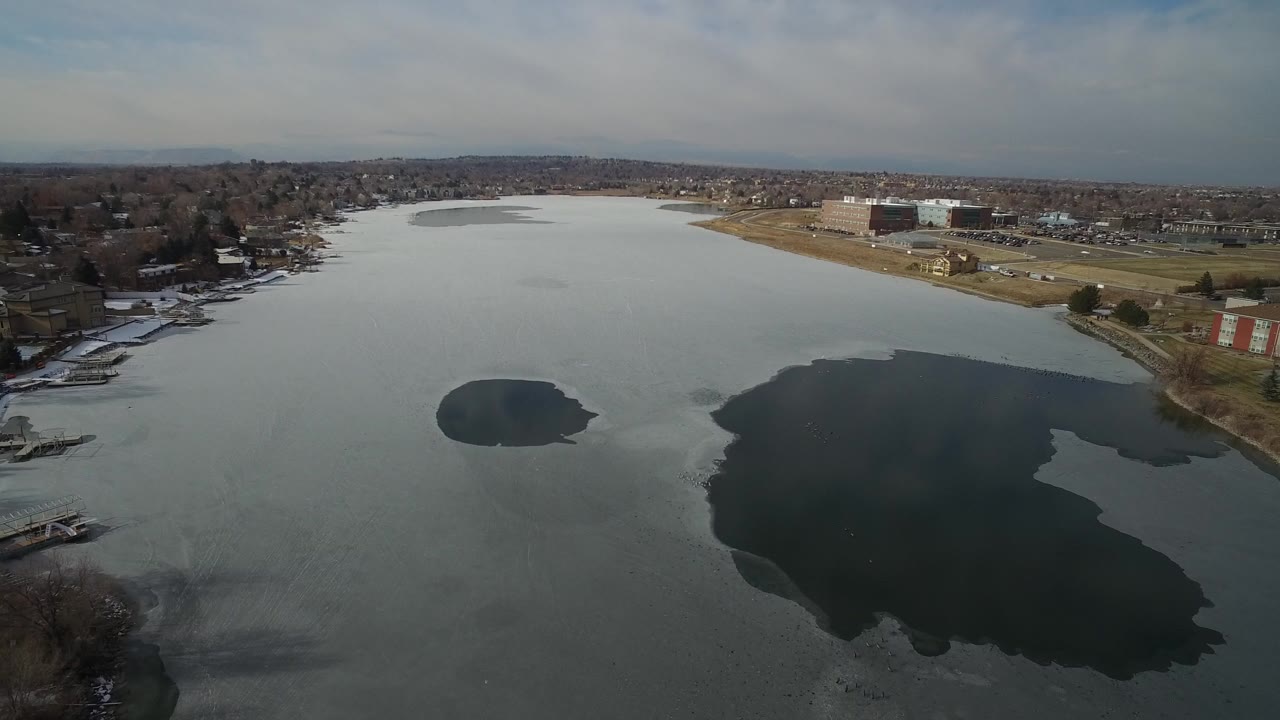 Aerial view of a frozen lake with dark open water patches and shoreline homes.