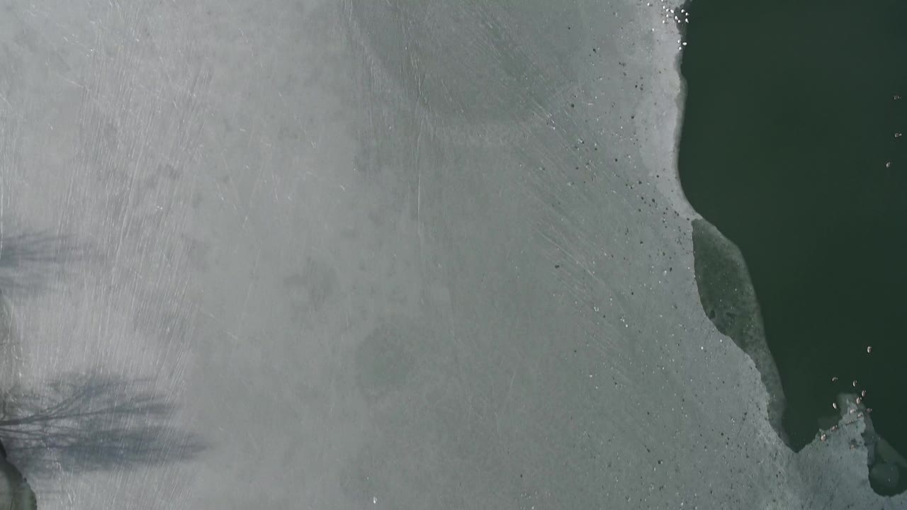 A near-vertical aerial frame over Hidden Lake, Colorado, showing a narrow dark green water channel bordered by pale ice, small bright flecks, and no visible people.
