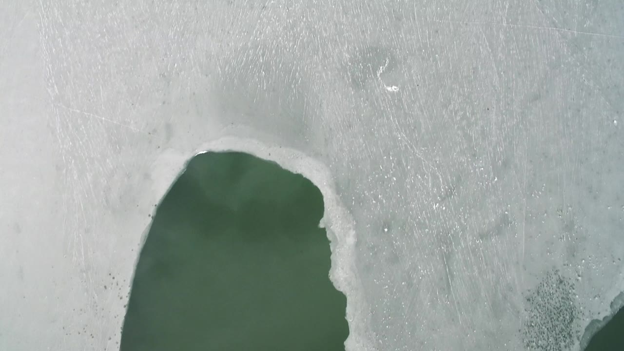 At 0:01, the aerial frame centers pale snow and scratched ice texture while part of the green U-shaped water body remains near the lower edge, with no visible people.