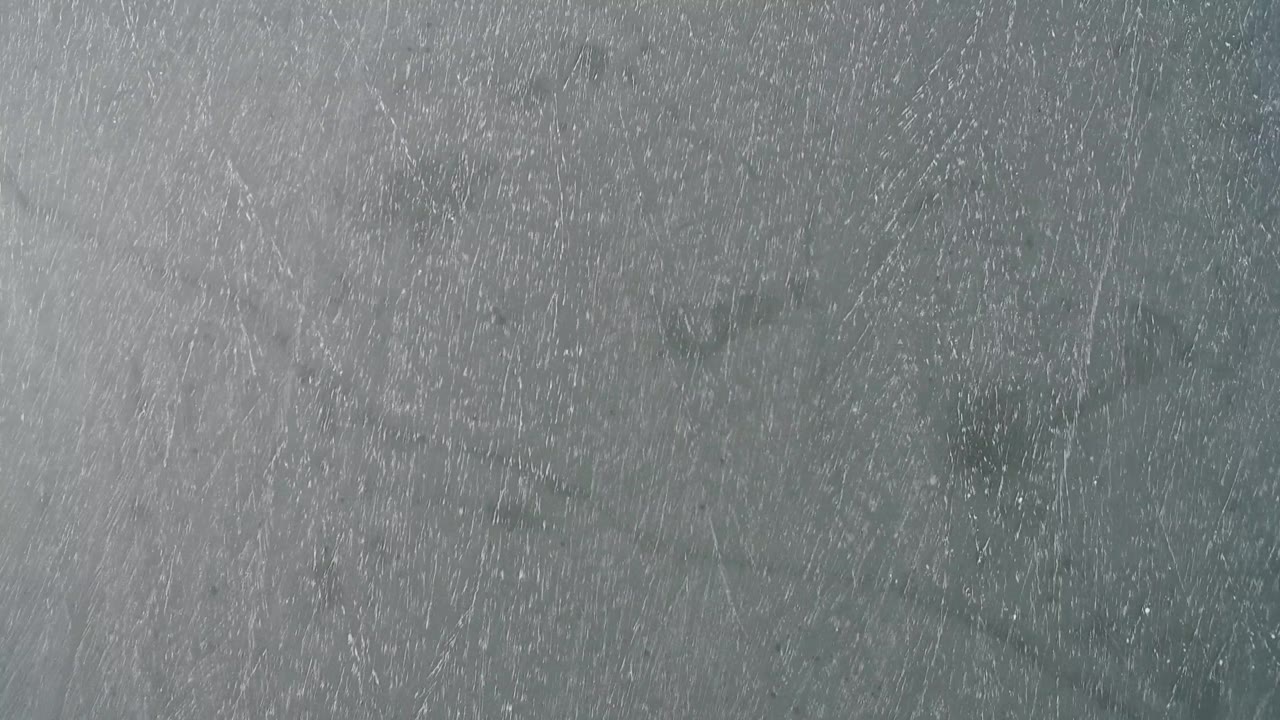 A close overhead frame shows pale gray-blue surface texture at Hidden Lake, Colorado, with darker shapes beneath and no visible people.