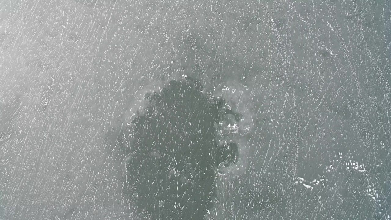 Overhead view of a gray-green frozen surface in Hidden Lake, Colorado, with dense white ice scratches, dark patches near the upper left, and no visible people.