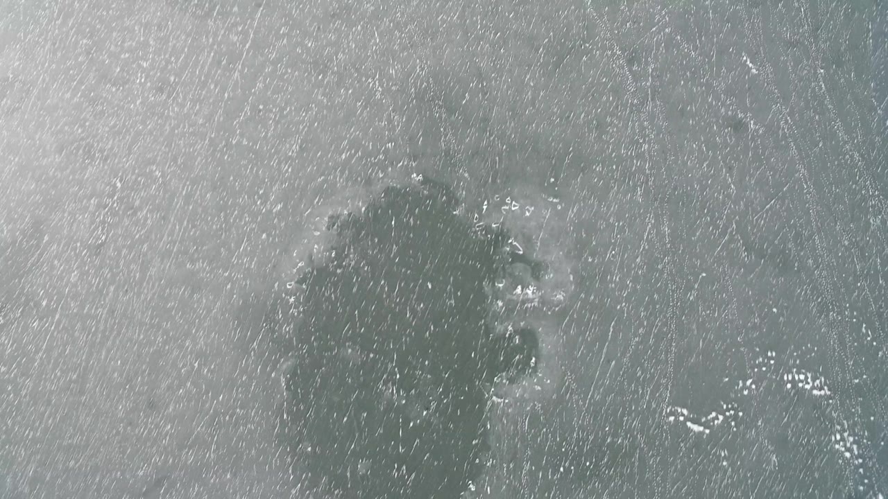 Overhead Hidden Lake, Colorado frame showing a scratched gray ice field with a dark irregular cluster near the upper center and no visible people.