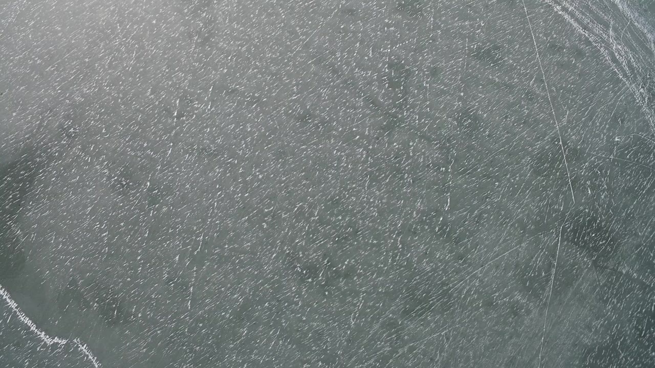 Overhead view of the frozen surface at Hidden Lake Colorado, showing gray green ice, white scratch patterns, long cracks, and no visible people.