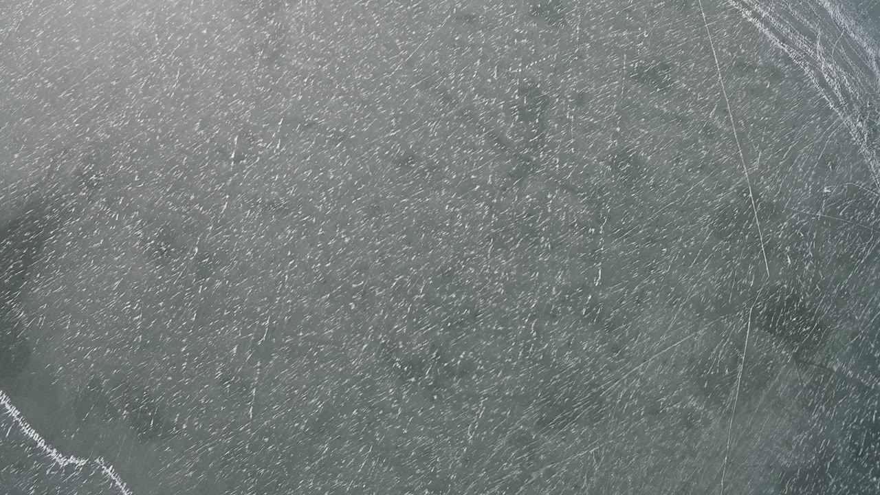 High overhead frame from Hidden Lake Colorado with dense white ice scratches, a darker oval patch near the center, faint linear cracks, and no visible people.