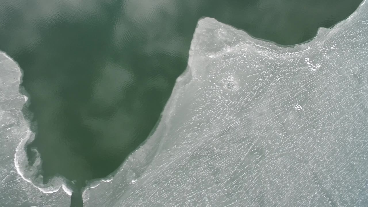 A curved pale ice margin crosses the frame over dark green water at Hidden Lake, Colorado, with cloudy surface markings and no visible people.