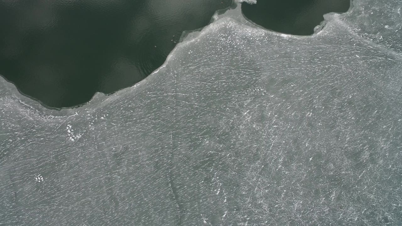 Hidden Lake, Colorado appears as a pale gray frozen sheet with fine crack lines and a rounded dark water opening near the top, with no visible people.
