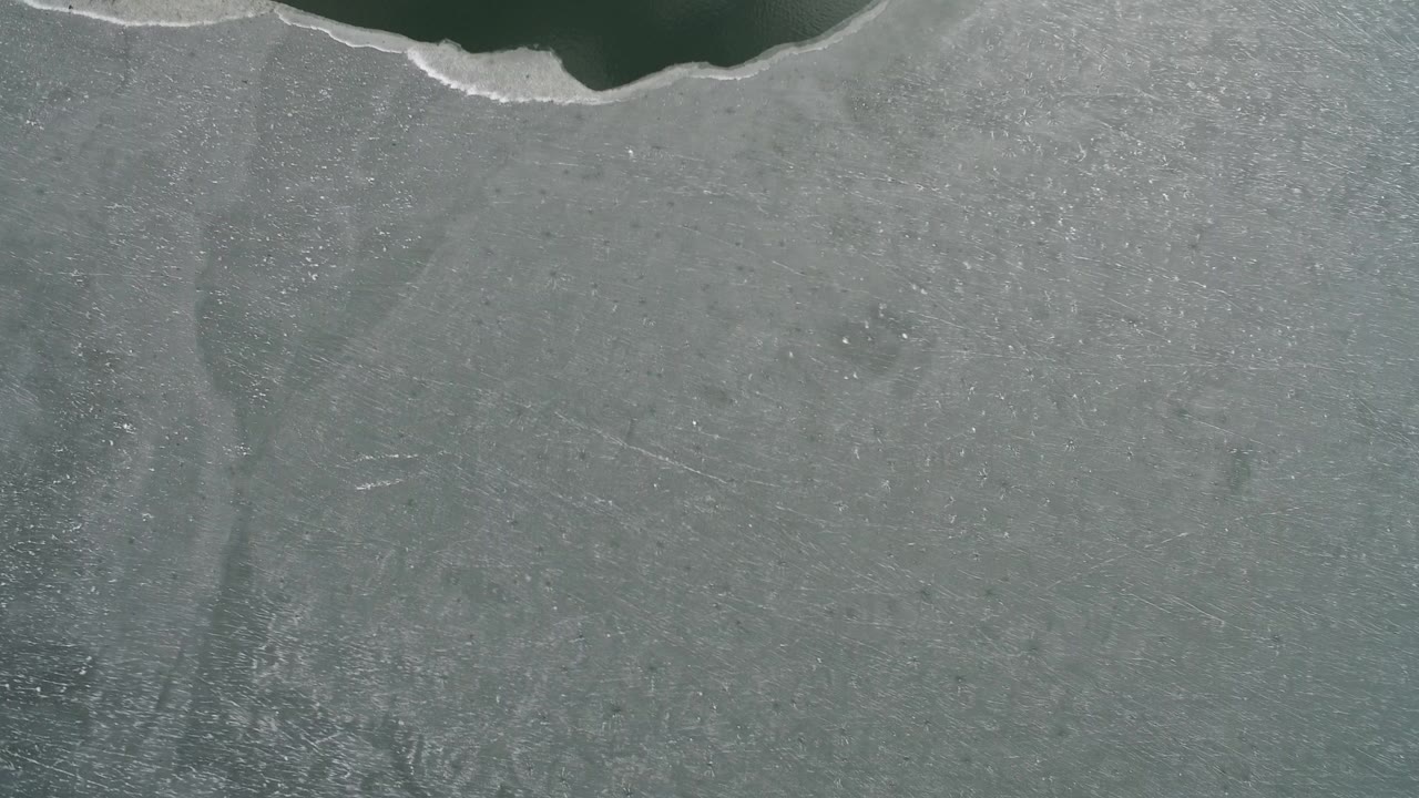 Overhead frame from Hidden Lake, Colorado showing gray textured lake ice below a narrow dark water edge, with no visible people.