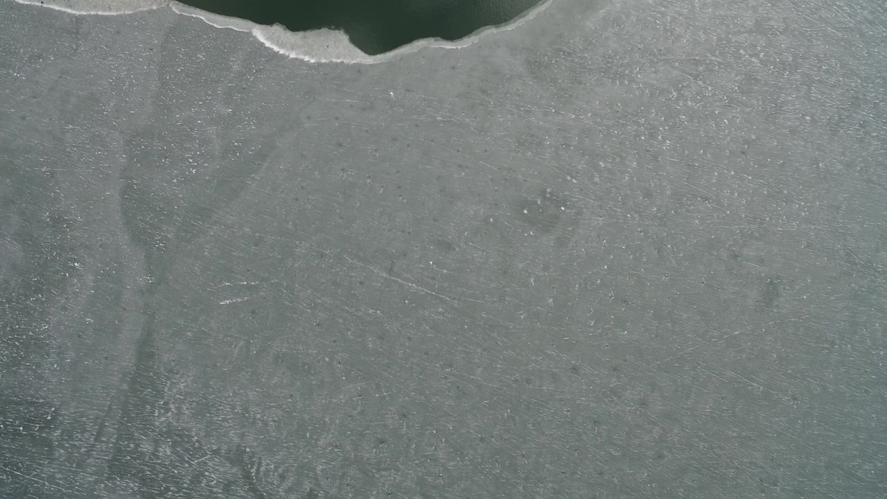 Hidden Lake, Colorado appears as a flat overhead field of gray-green ice with a scalloped dark water strip near the top and no visible people.