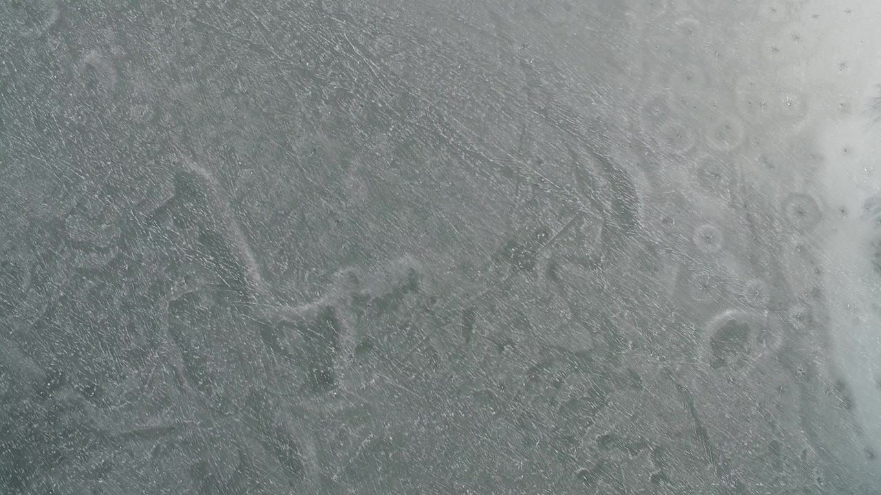 Top-down view of gray frozen surface at Hidden Lake, Colorado, with pale snow-like streaks, circular bubble marks, and no visible people.