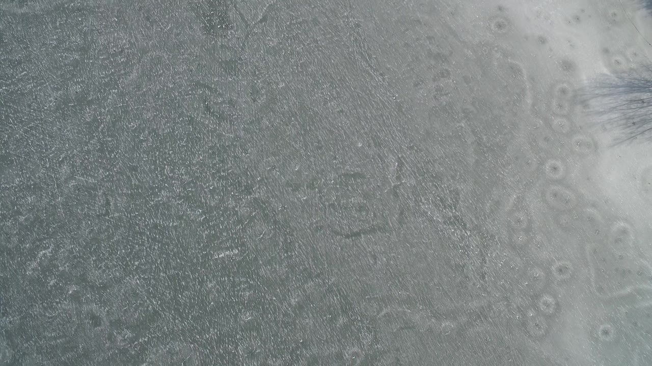Close aerial frame over Hidden Lake, Colorado showing scratched gray ice, pale cloudy ice areas, and no visible people.