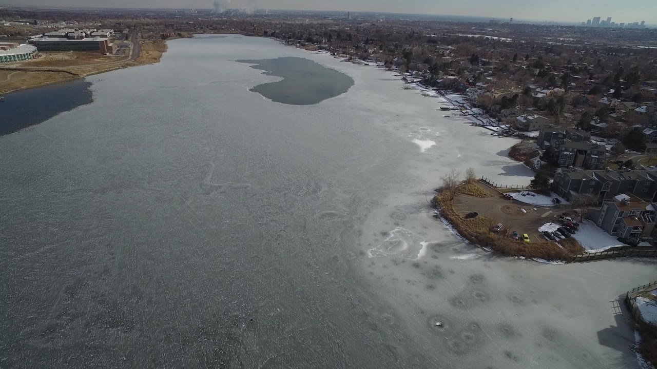 Aerial view over Hidden Lake, Colorado, with gray frozen water in the foreground, snow along the right shoreline, homes beyond the ice, and no visible people.