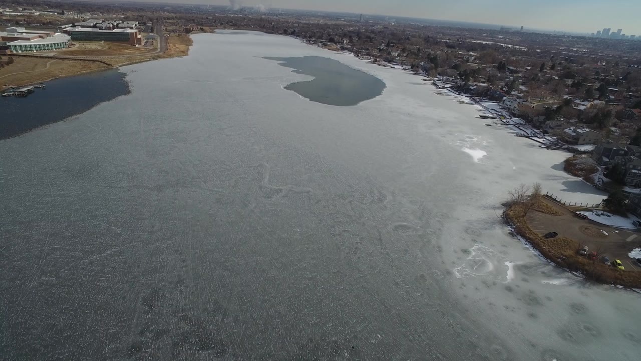 Opening aerial frame over Hidden Lake, Colorado, with dark open water at left, pale thawing ice across the center, homes along the right shoreline, and no visible people.