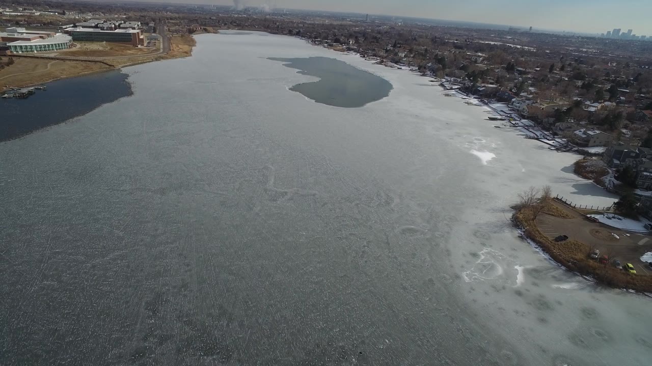 Aerial view over Hidden Lake, Colorado, showing a wide pale ice field, a darker open-water inlet on the left bank, lakefront houses on the right, and no visible people.
