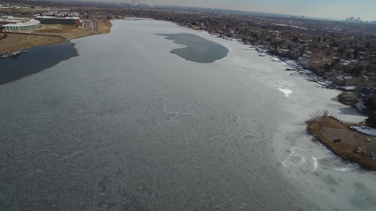Aerial lake view with a dark open-water patch and distant town skyline.
