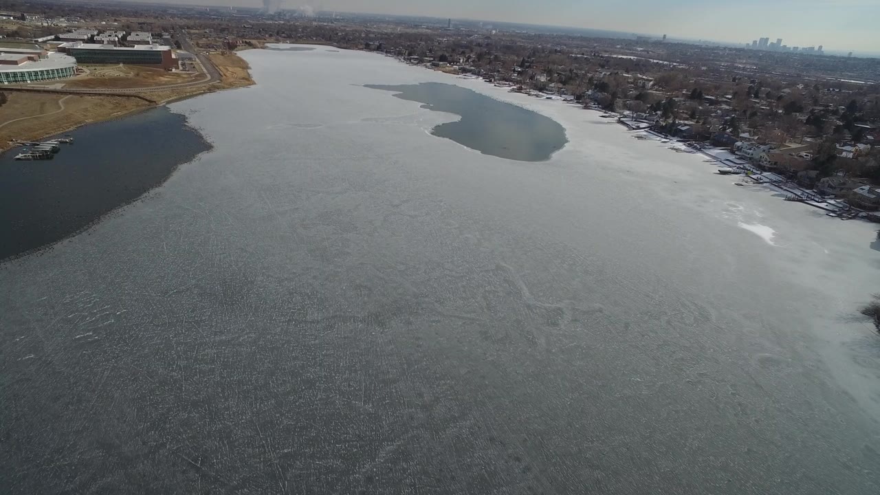 High aerial frame over Hidden Lake, Colorado showing gray water or ice texture in the foreground, shoreline homes in the distance, a large complex at left, and no visible people.