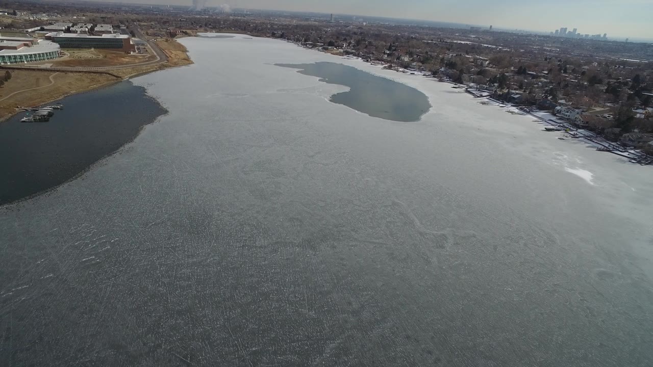 Drone view across a gray body of water toward houses and a distant city skyline.