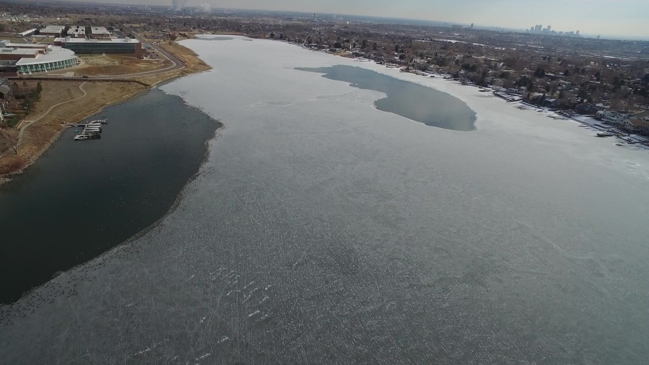 Aerial frame over Hidden Lake, Colorado showing dark open water at left, pale ice across the lake or river, brown shoreline, large buildings, residential blocks, and no visible people.