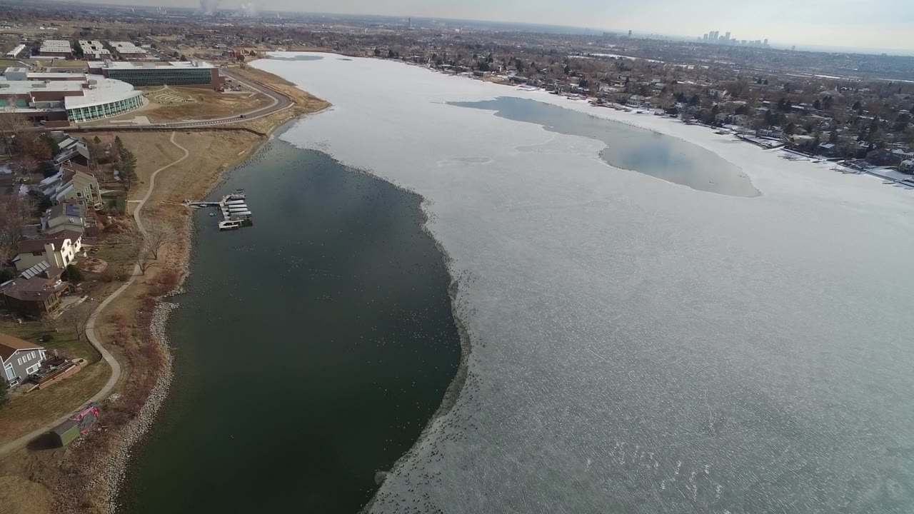 High view over Hidden Lake, Colorado showing the curved open-water channel beside shore homes, small docks, broad lake ice, and no visible people.