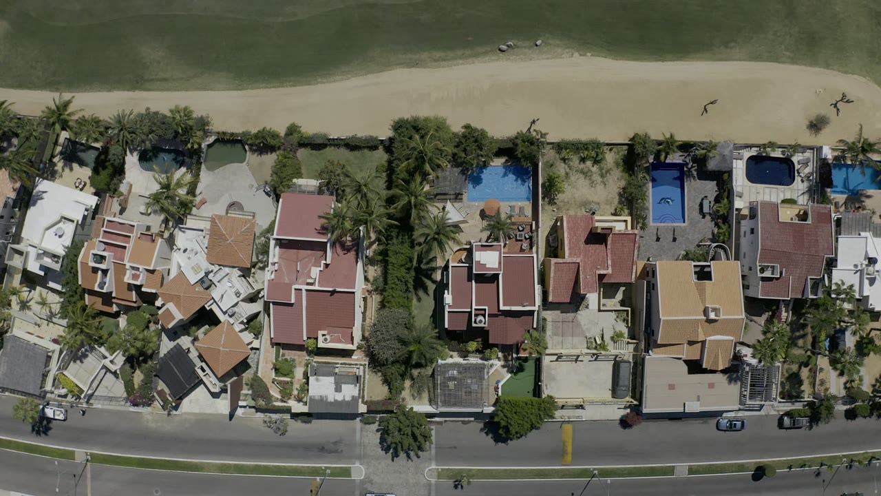A Los Cabos overhead frame centers a row of private houses and blue pools between a curved street below and a narrow beach with green water above, with tiny people visible near the shore.