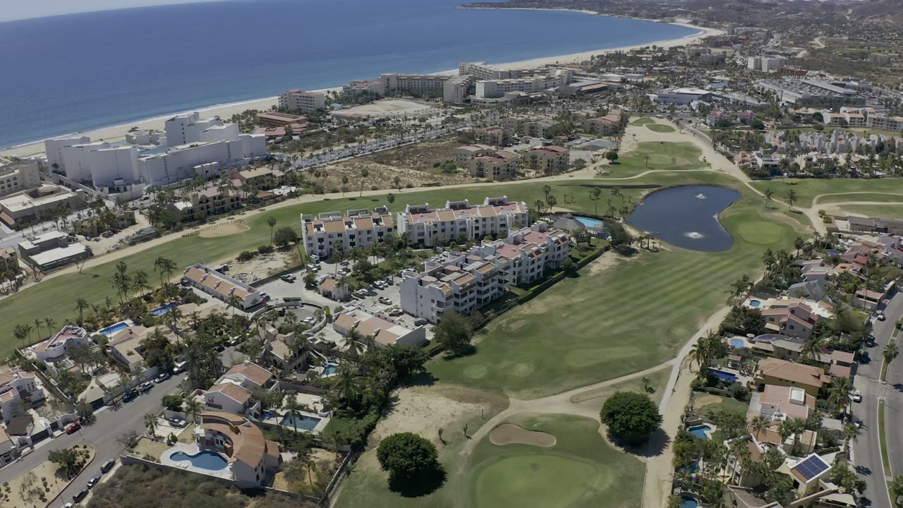 Drone view in Los Cabos with condo blocks centered beside green fairways, curving residential streets, a pond to the right, and the beach and ocean beyond, with no visible people.