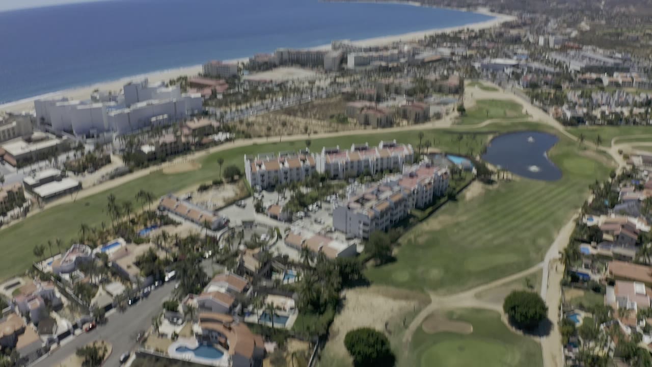 Aerial frame in Los Cabos, Mexico, with a blurred oblique view of homes, fairways, streets, a pond, and the distant beach and ocean.