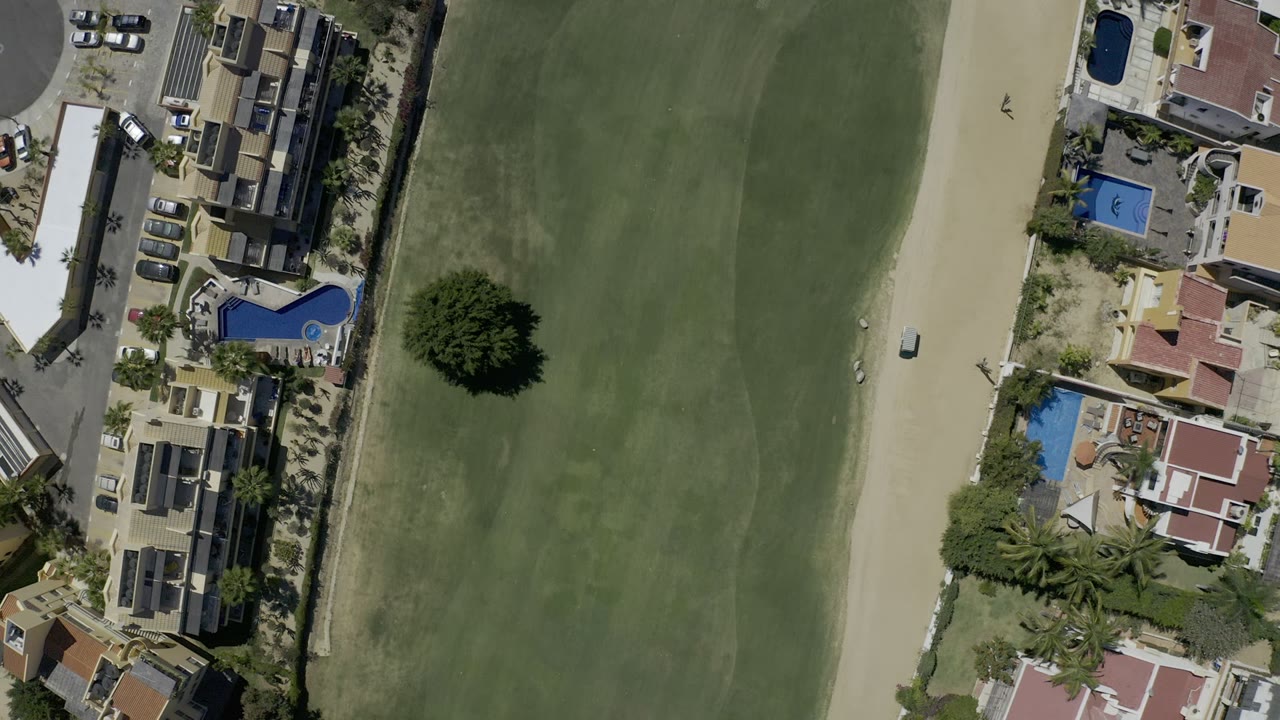 Opening overhead frame in Los Cabos, Mexico showing green grassland beside a sand path, private homes, blue pools, parked cars, palms, and no visible people.