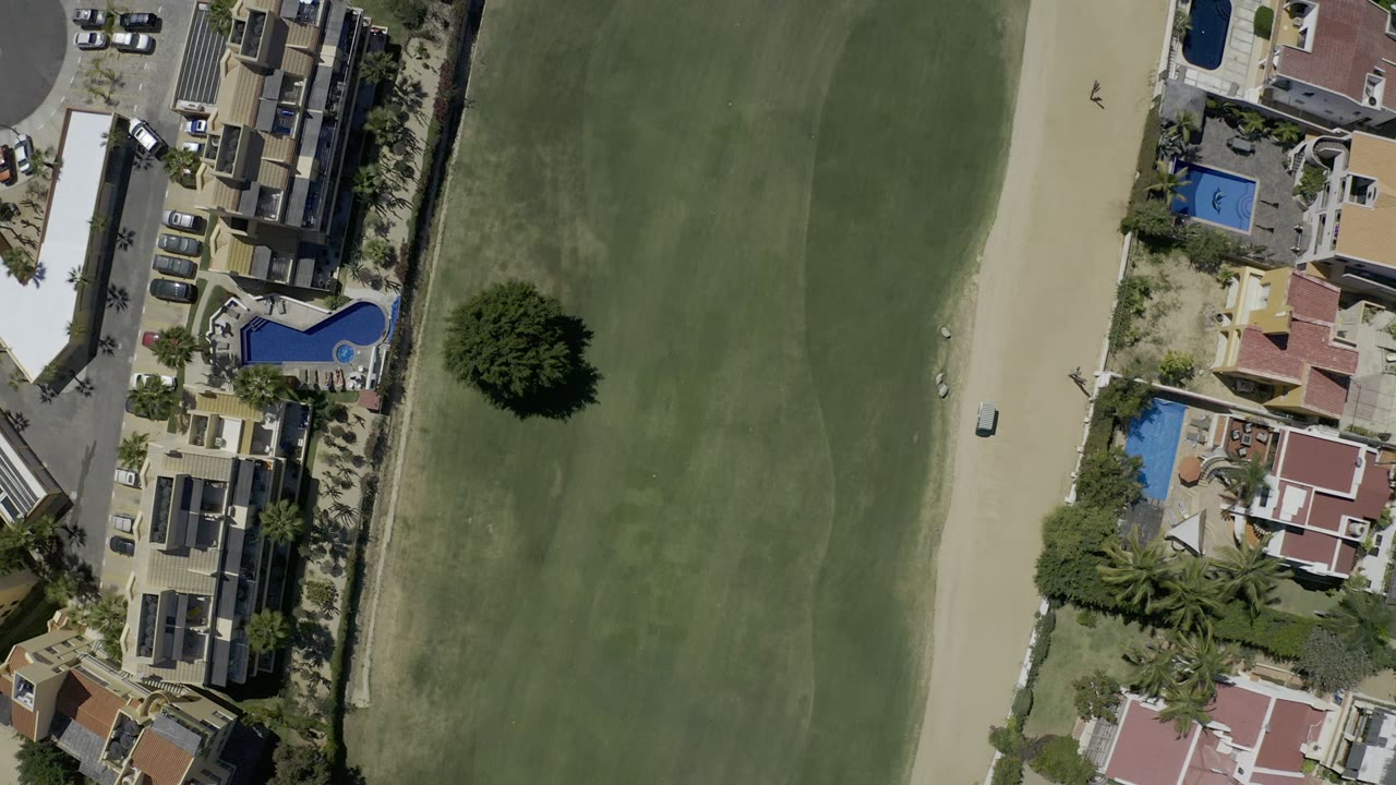 A small white vehicle appears on a pale sand road beside green grassland and homes.