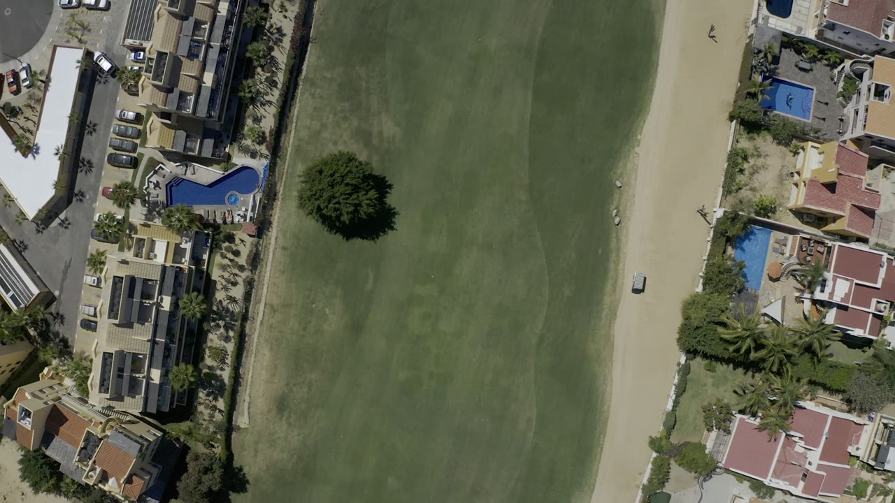 Mid-clip overhead frame in Los Cabos, Mexico showing the white vehicle farther along the sand road, bordered by green grassland, homes, pools, trees, and no visible people.