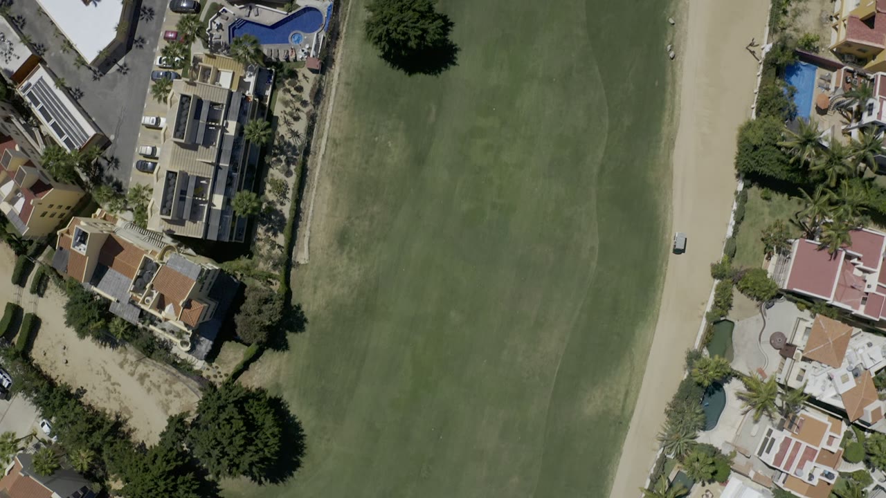 Overhead view of green fairway, pale path, houses, trees, and parked cars.