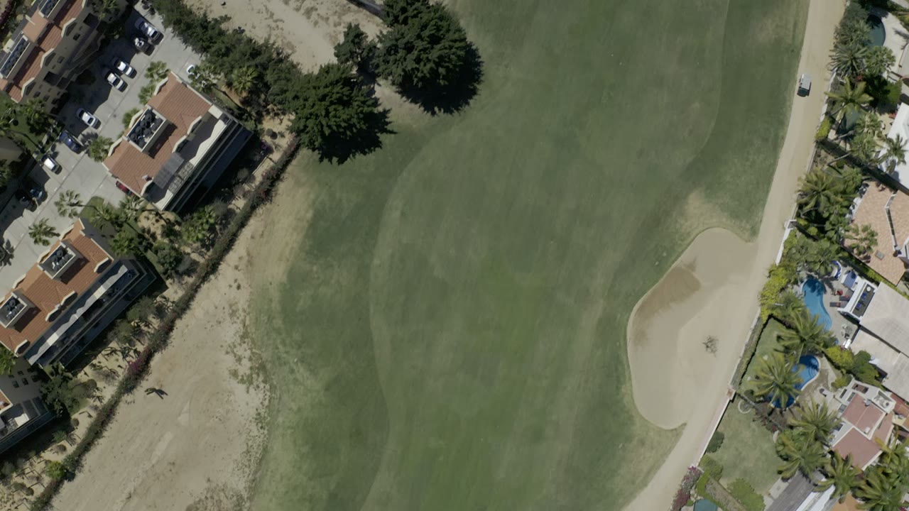 Top-down aerial frame of a Los Cabos golf course with green fairway, pale sand bunker, cart path, tile-roof buildings, palms, and no visible people.