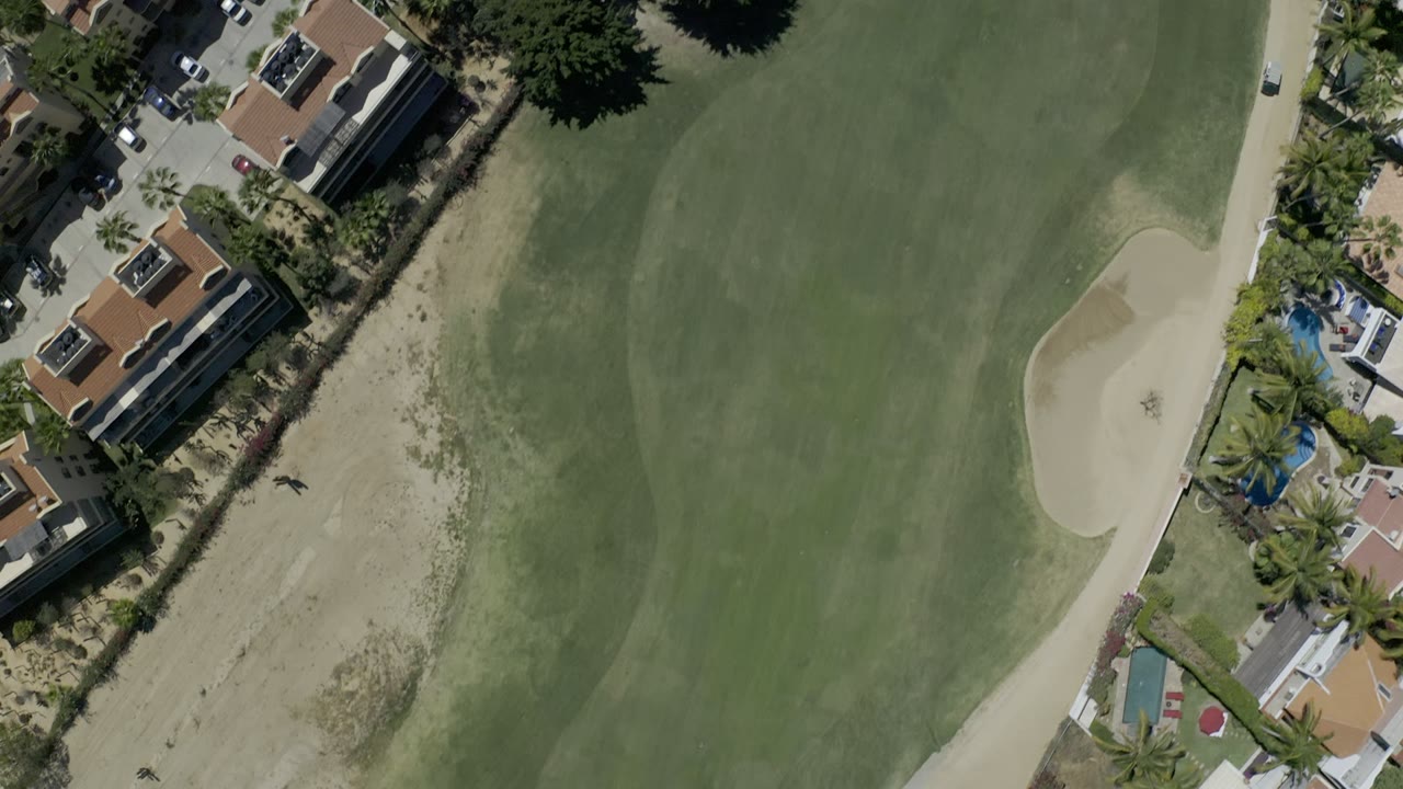 Aerial Los Cabos golf frame with broad green fairway, rough sandy ground, a curved bunker edge, nearby residential buildings, palms, and no visible people.