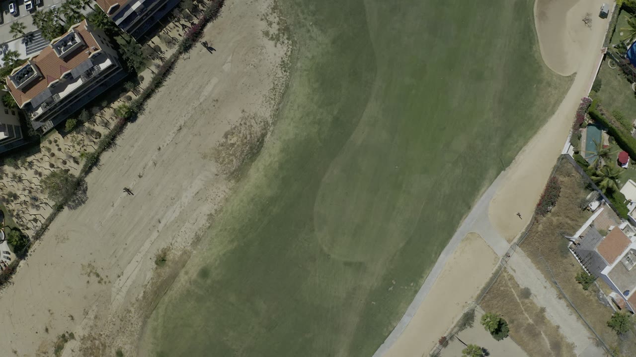 Overhead Los Cabos frame with the fairway shifted across the image, pale sand along one side, a cart path at the edge, homes, and small human figures visible on the sand.