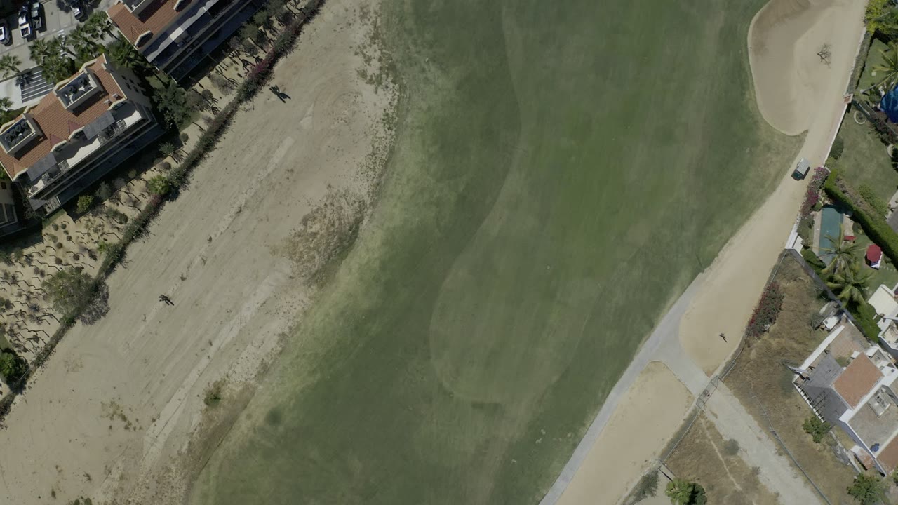 Top-down aerial view of green grass, sandy rough, homes, and a curved path.