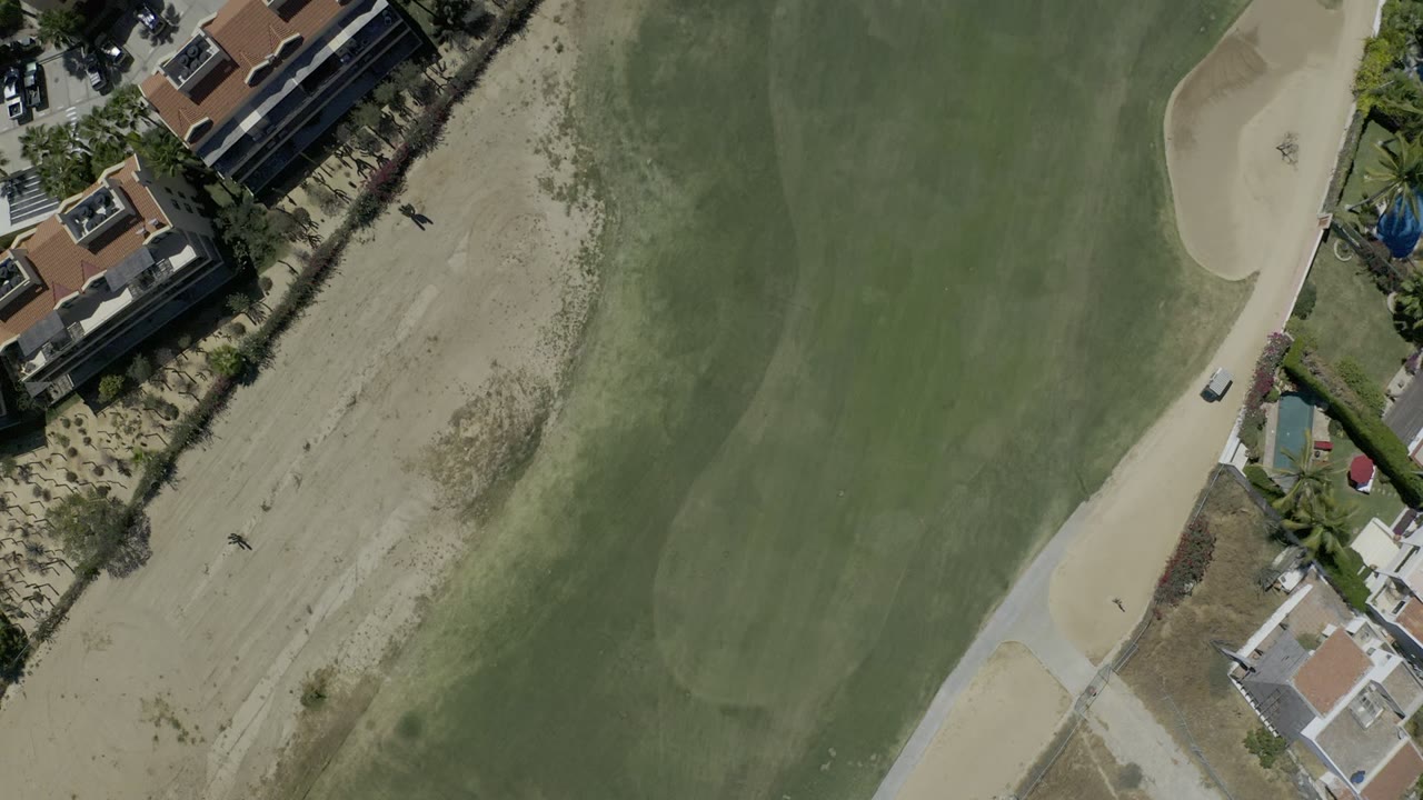 High overhead Los Cabos frame with a sand bunker near the upper edge, broad green turf, roadside homes, palms, and no visible people.