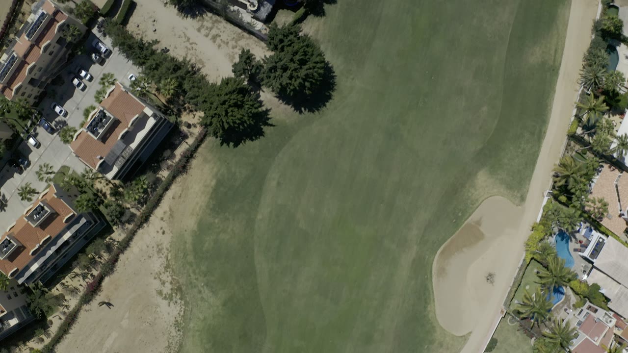 Overhead view of golf-course grass beside houses, trees, sandy ground, and a small distant figure.