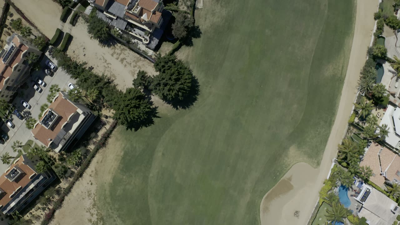 Aerial Los Cabos frame with a broad green fairway, pale sandy border, private homes, palm trees, parked cars, and a blue pool visible near the residential lots.