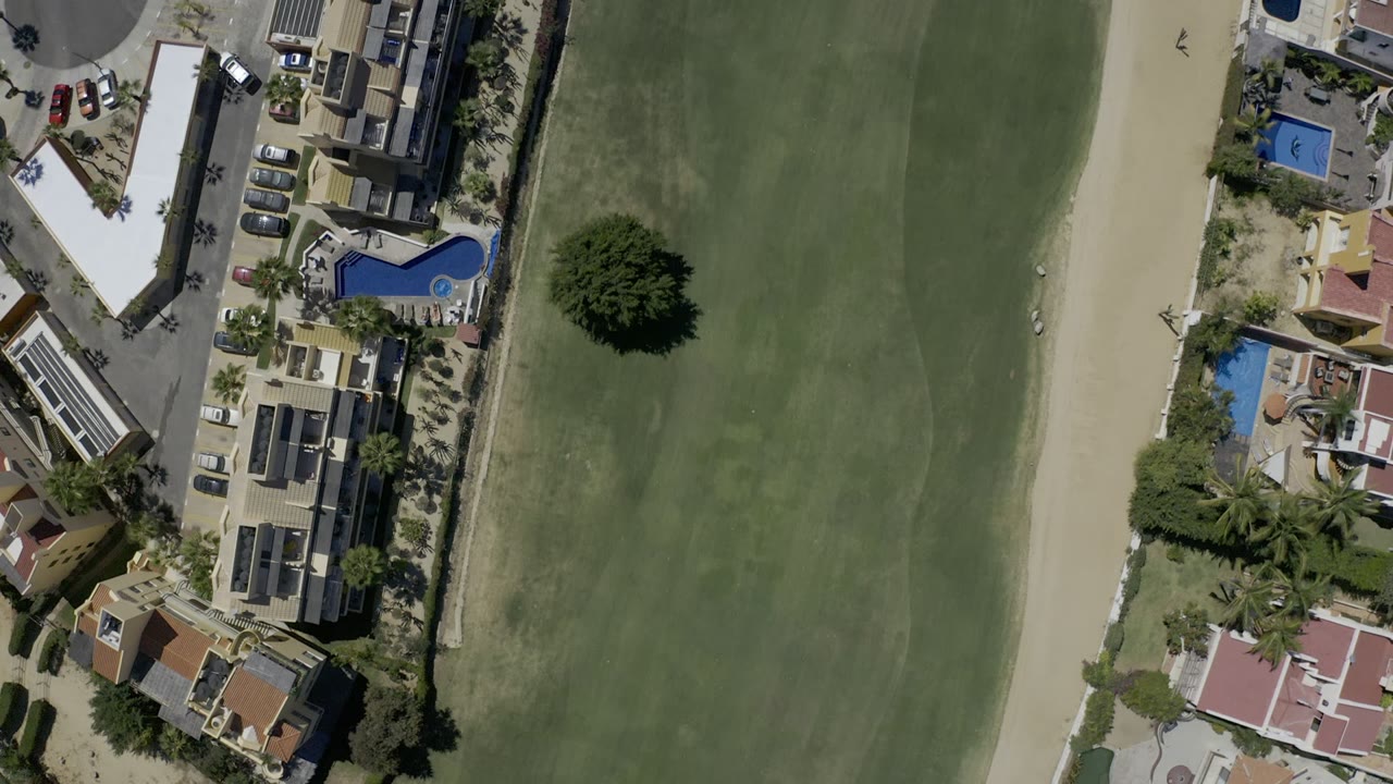 One second in, the Los Cabos aerial view places the green lawn through the center with a curved blue pool, residential roofs, palm trees, parked cars, and no visible people along the edge.