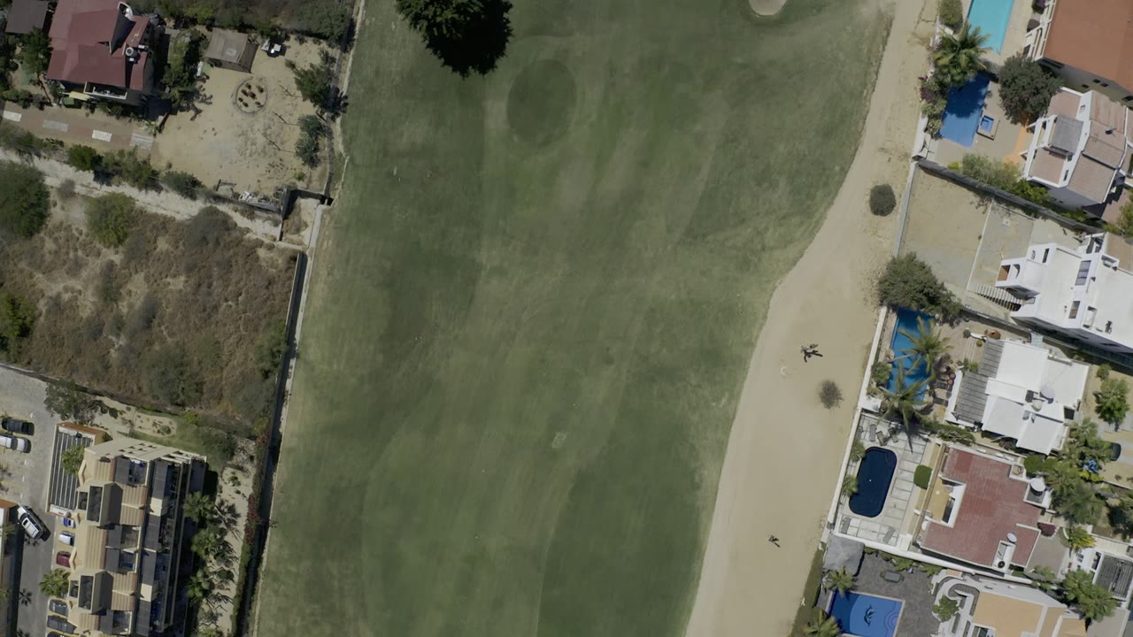 Overhead Los Cabos golf course frame with the fairway filling the left side and private houses, pool areas, walls, and dry lots along the right edge, with no visible people.