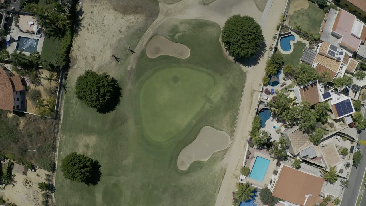 A later overhead frame in Los Cabos, Mexico shows the fairway edge, putting green, light sand bunkers, clustered homes, several pools, and no visible people.